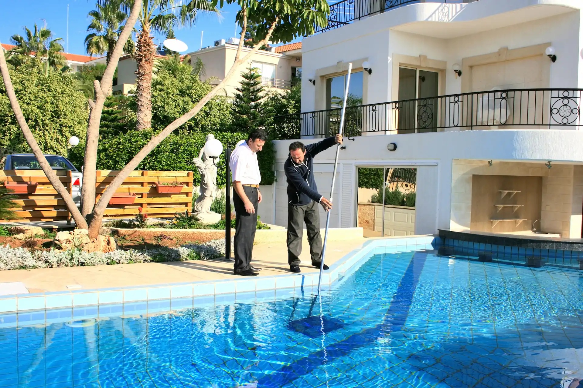 Medium shot of a pool maintenance worker vacuuming an inground pool as a pool owner observes. A pool maintenance service like this is one way to make pool customers' lives easier.