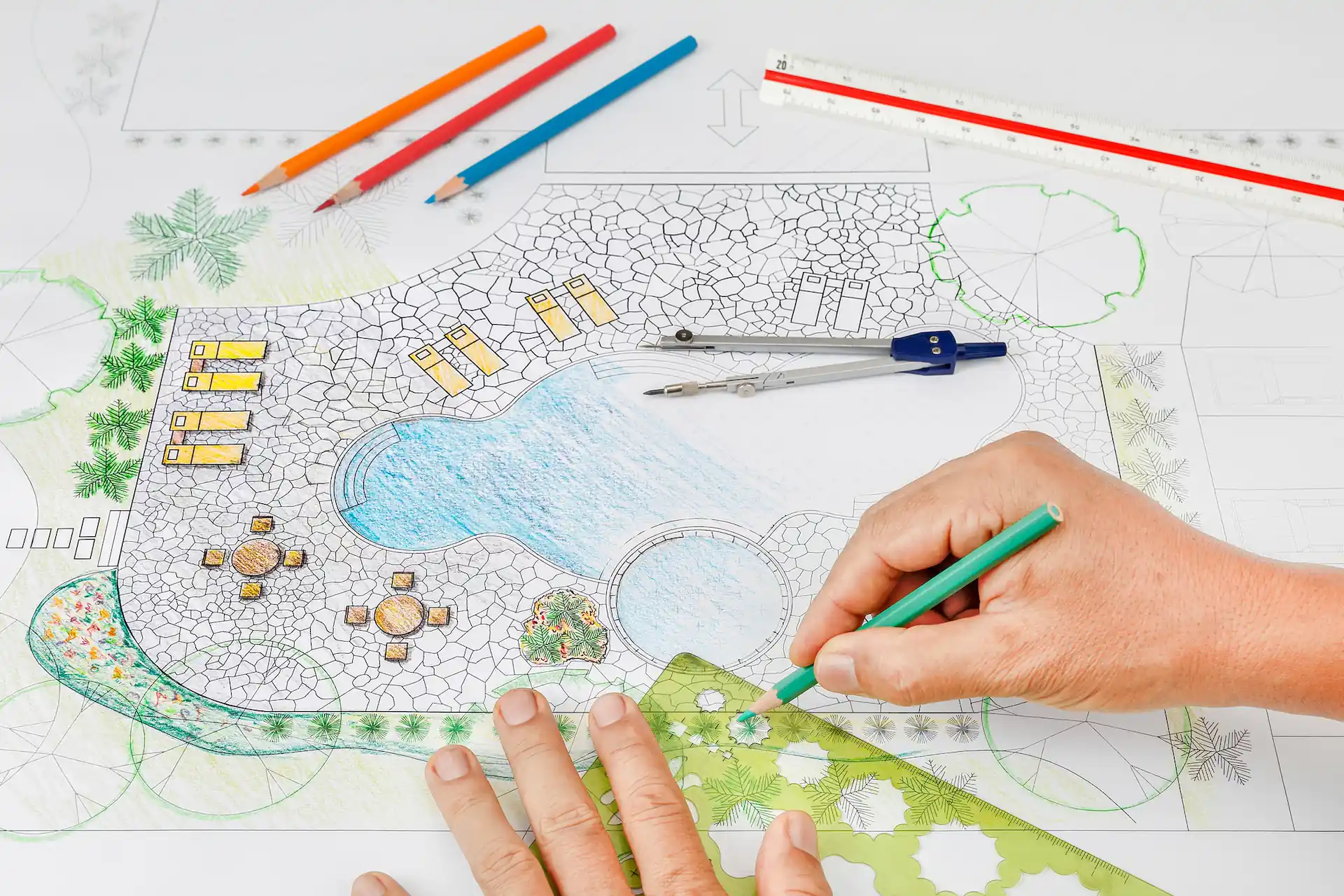 Overhead shot of a person drawing an elaborate pool and backyard design with colored pencils on draft paper. It takes significant expertise and resources to add design services like these to a pool builder business.