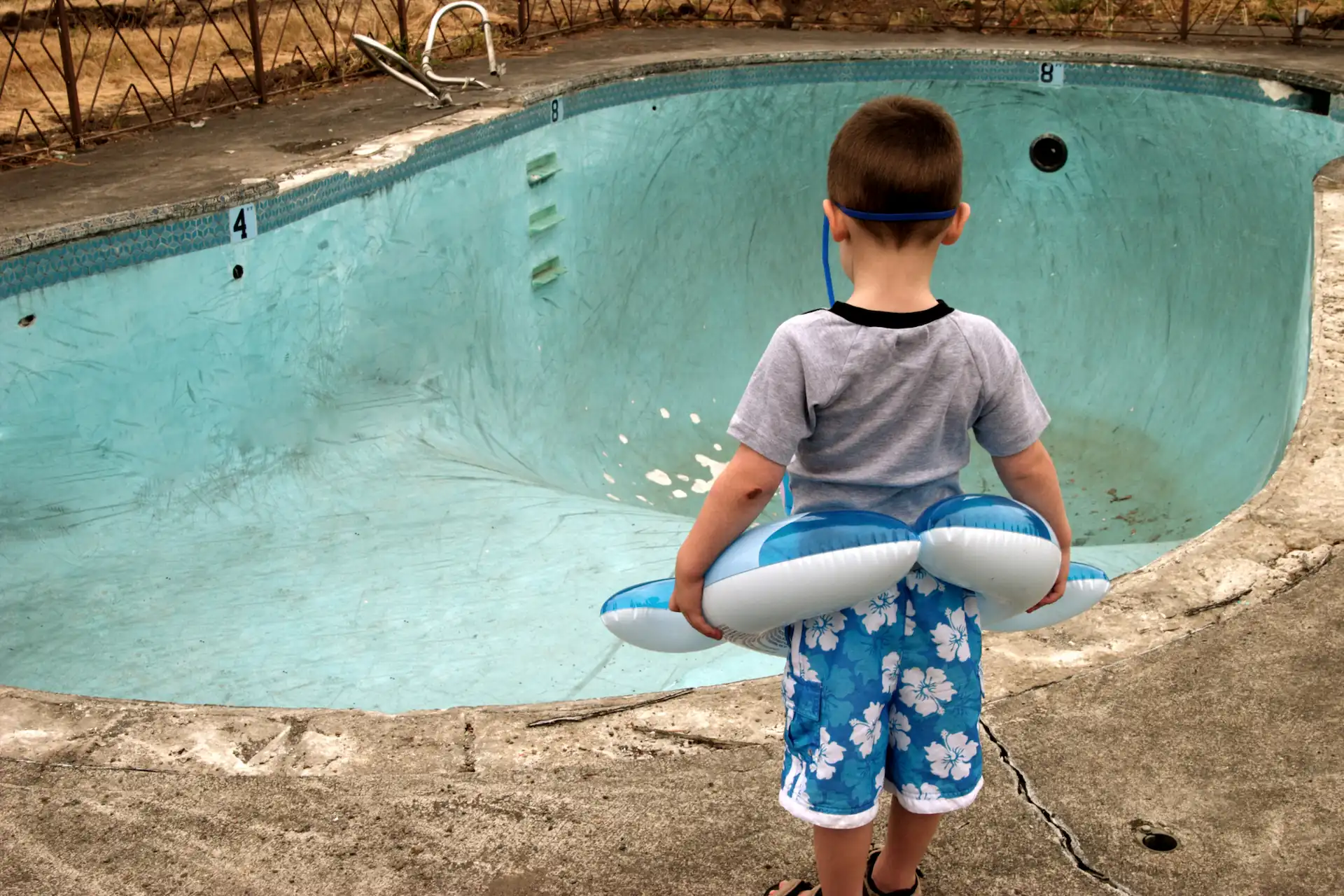 Medium shot of a young swimmer standing at the edge of an empty, dirty in-ground pool while holding a floatie. When pool builders aren't generating pool leads, the business can feel as bleak and dry as this scene.