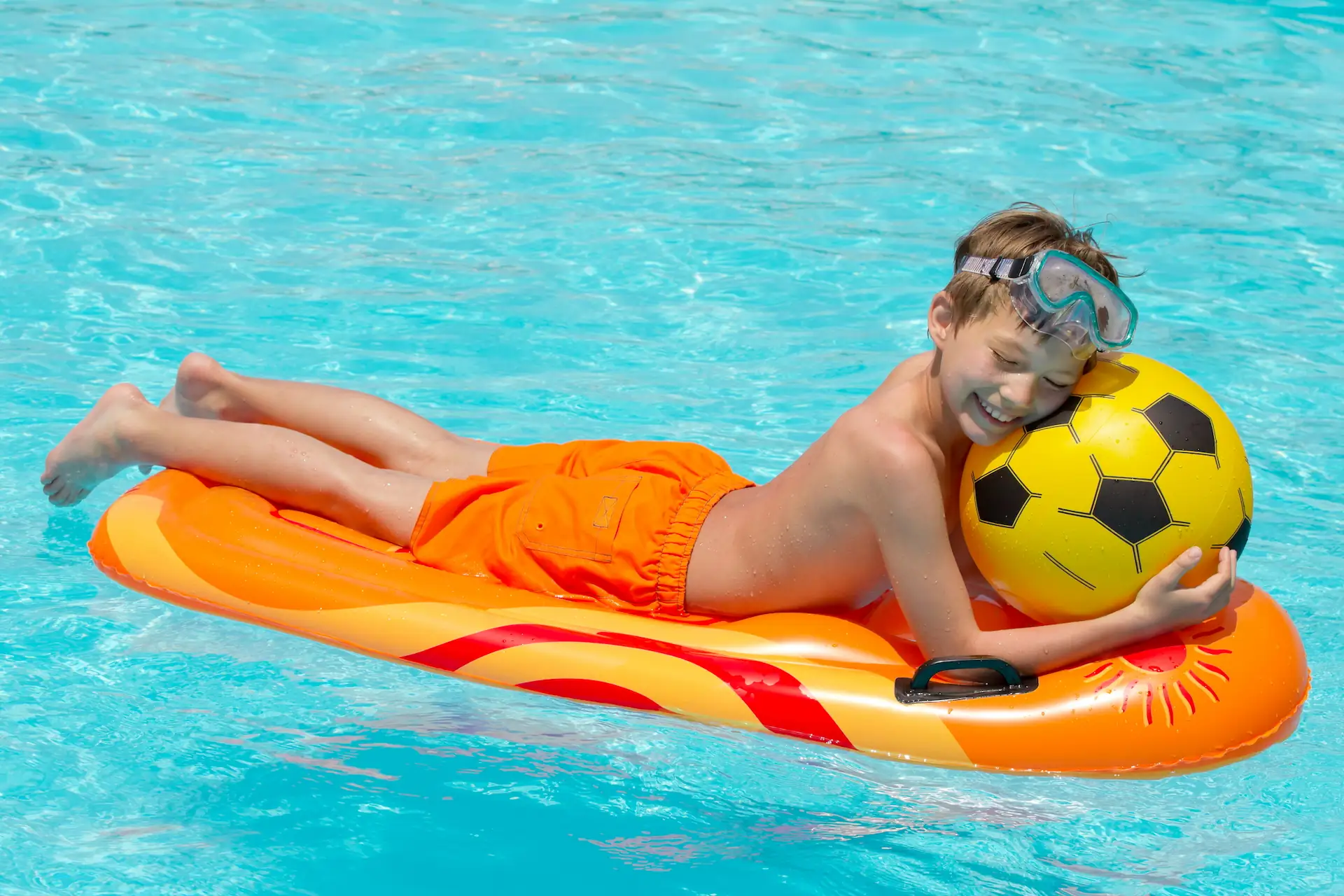 Medium shot of a young swimmer in orange swim trunks laying face down on an orange pool float while embracing a beach ball that looks like a yellow soccer ball. It's important the pool products like floats and toys be high-quality for the sake of user safety and enjoyment.