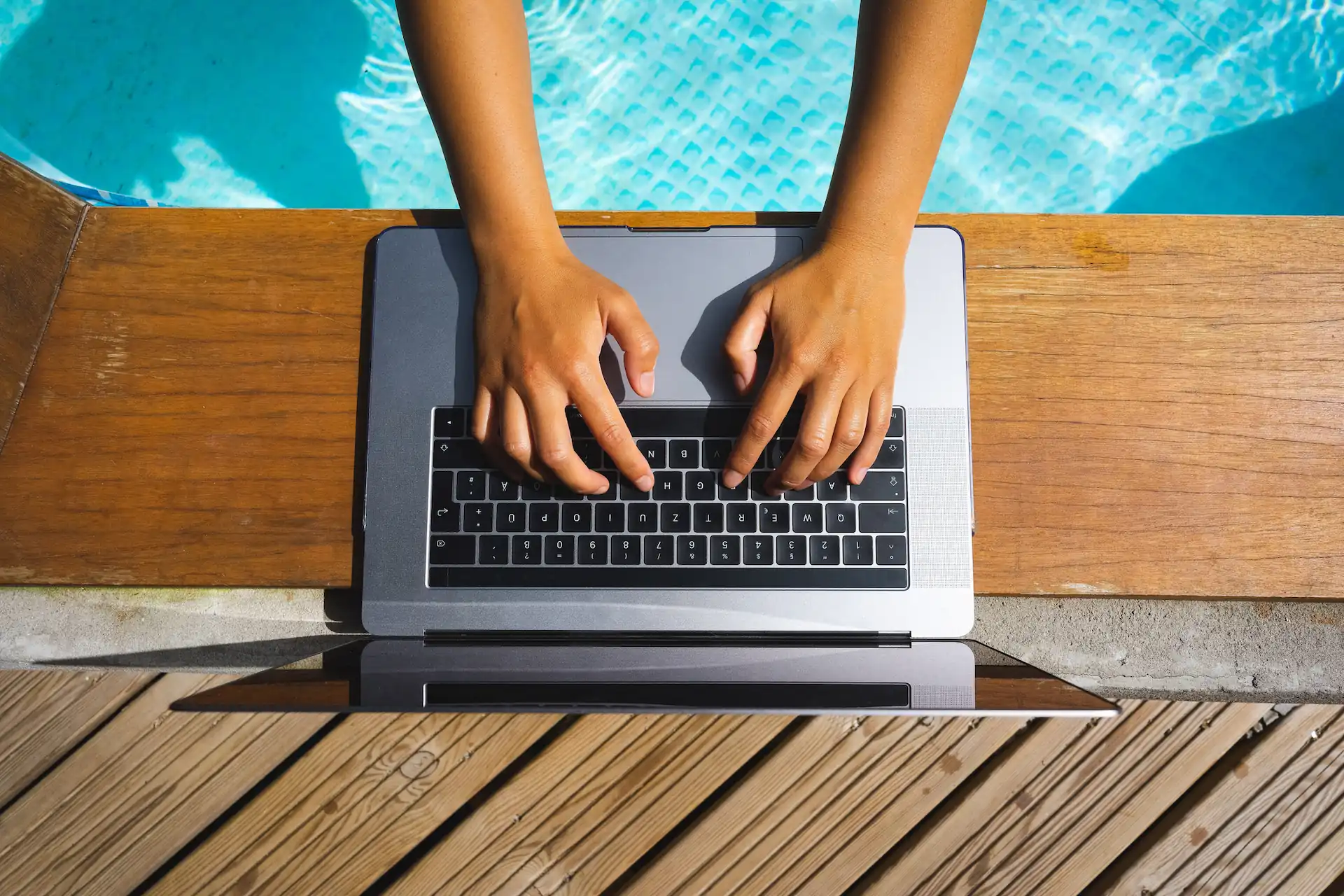 Overhead shot of a person's hands typing on a laptop keyboard placed at the edge of an inground pool. Part of maintaining a solid online presence for a pool business is dealing with online reviews.