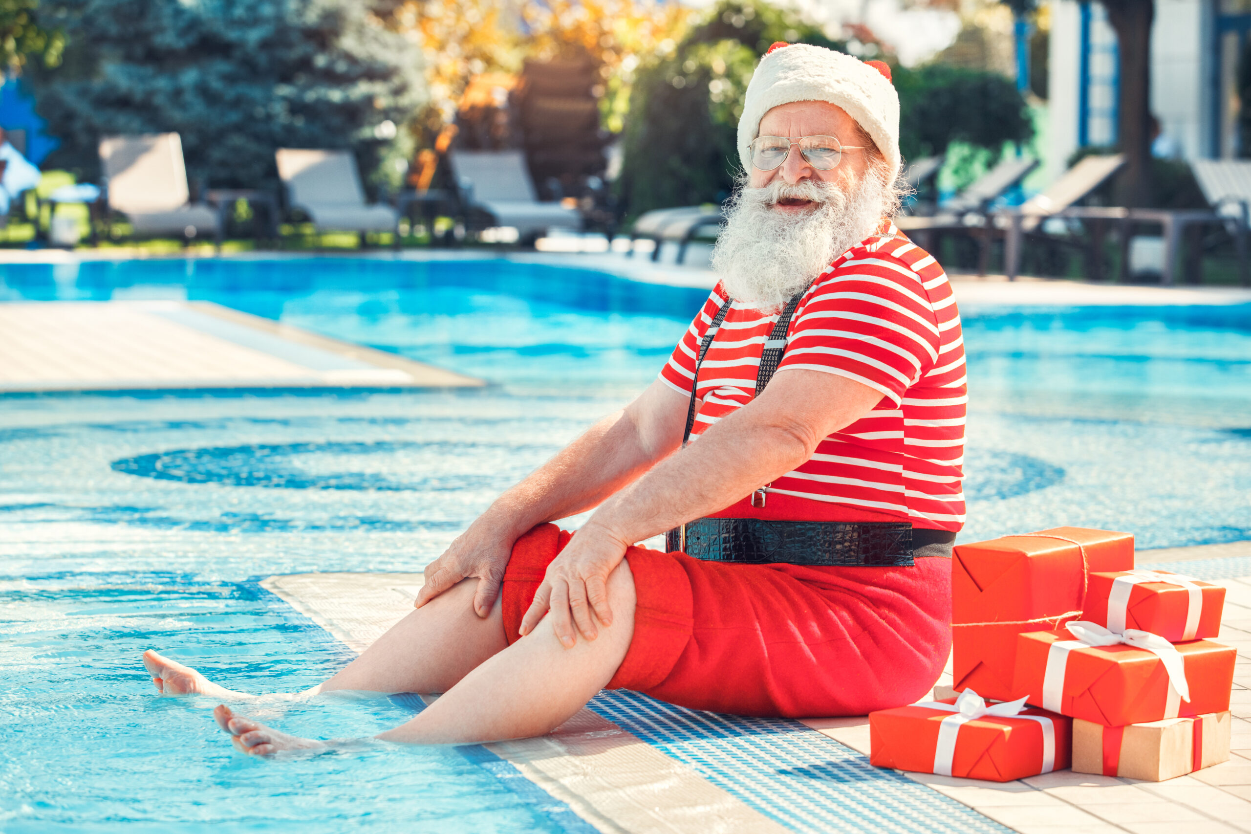 A man in a Santa outfit is sitting on the edge of a pool with his feet in the water. There are red presents next to him.